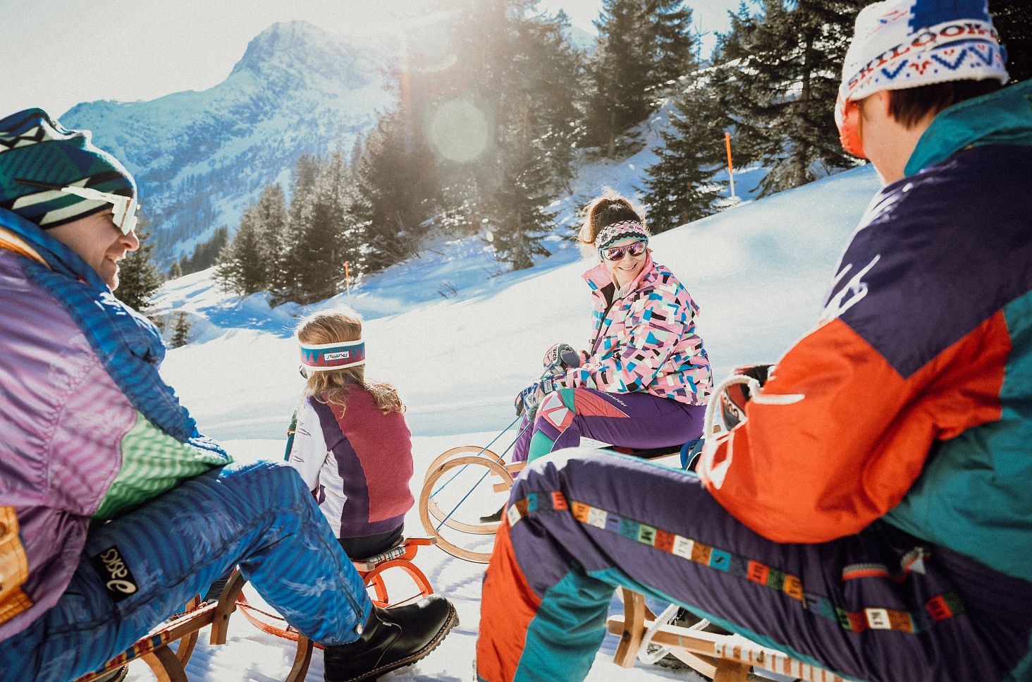 Vier Personen sitzen mit Holzschlitten im Schnee und blicken auf die verschneite Berglandschaft.