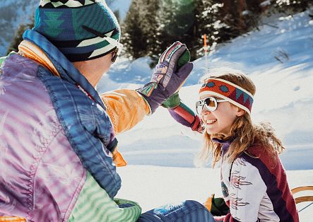 Eine erwachsene Person und ein Kind sitzen im Schnee und geben sich mit den Handschuhen ein High Five.