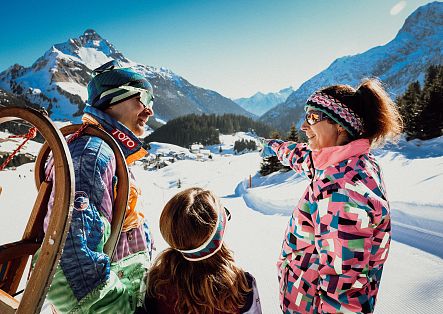 Drei Personen mit Schlitten stehen im Schnee und betrachten gemeinsam die umliegenden verschneiten Berge.