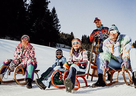Personen sitzen auf einem Holzschlitten und fahren durch eine verschneite Winterlandschaft.
