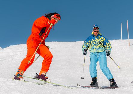 Zwei Skifahrer stehen auf einer Piste, während einer von ihnen einen dynamischen Sprung ausführt.