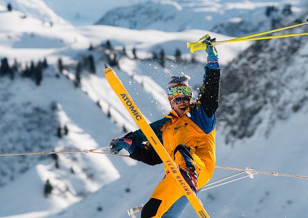 Ein Skifahrer springt mit überkreuzten Skiern durch die Luft und hält dabei seine Skistöcke nach oben.