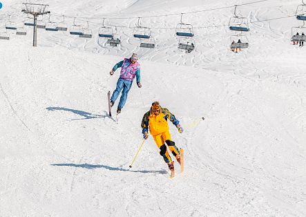 Zwei Skifahrende springen während der Abfahrt über eine verschneite Piste.