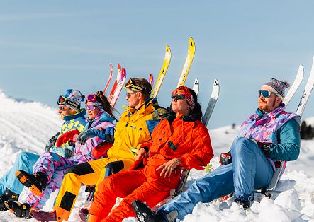 Fünf Skifahrende sitzen nebeneinander im Schnee und halten ihre Skier aufrecht hinter sich.