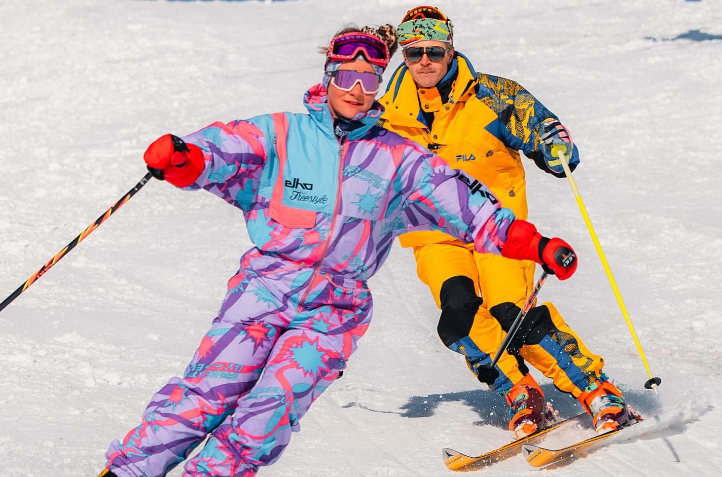 Zwei Skifahrende in farbenfroher Kleidung fahren gemeinsam eine verschneite Piste hinunter.