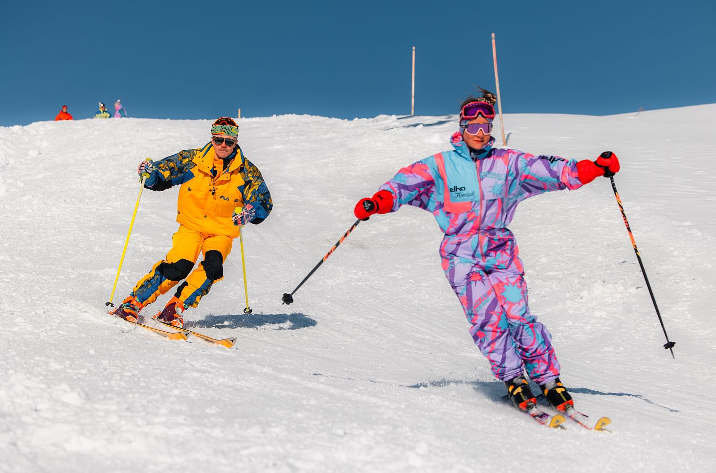 Two men in colourful 1980s ski suits are skiing down the slope side by side.