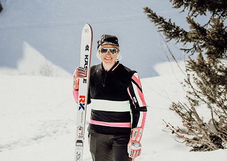 Eine Skifahrerin steht mit Skiern und Skistöcken vor einer verschneiten Berglandschaft.
