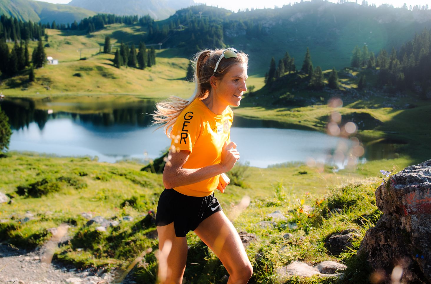A female trail runner in a yellow shirt is running along the shore of Lake Körbersee.