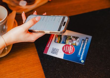 Eine Hand hält ein Smartphone über einem Holztisch, auf dem eine Informationskarte mit QR-Code liegt.