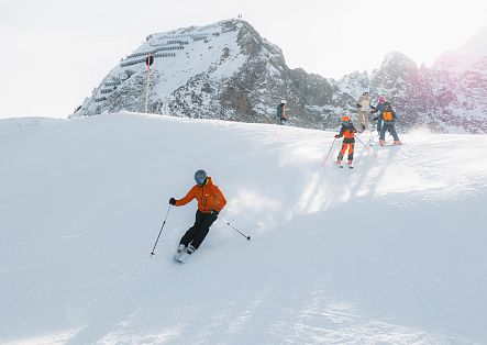 Eine Person in orangefarbener Skibekleidung fährt durch eine verschneite Landschaft, während weitere Skifahrende im Hintergrund zu sehen sind.