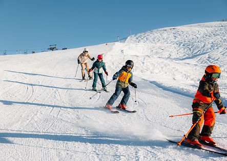 Several skiers are skiing down a snow-covered slope one after the other, whilst white mountains can be seen in the background.