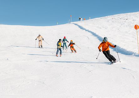 Mehrere Personen fahren auf einer breiten, sonnigen Skipiste bergab. Im Vordergrund fährt eine Person in orangefarbener Skijacke mit weit auseinanderstehenden Skiern und Skistöcken eine Kurve. Dahinter sind weitere Erwachsene und Kinder beim Skifahren zu sehen.