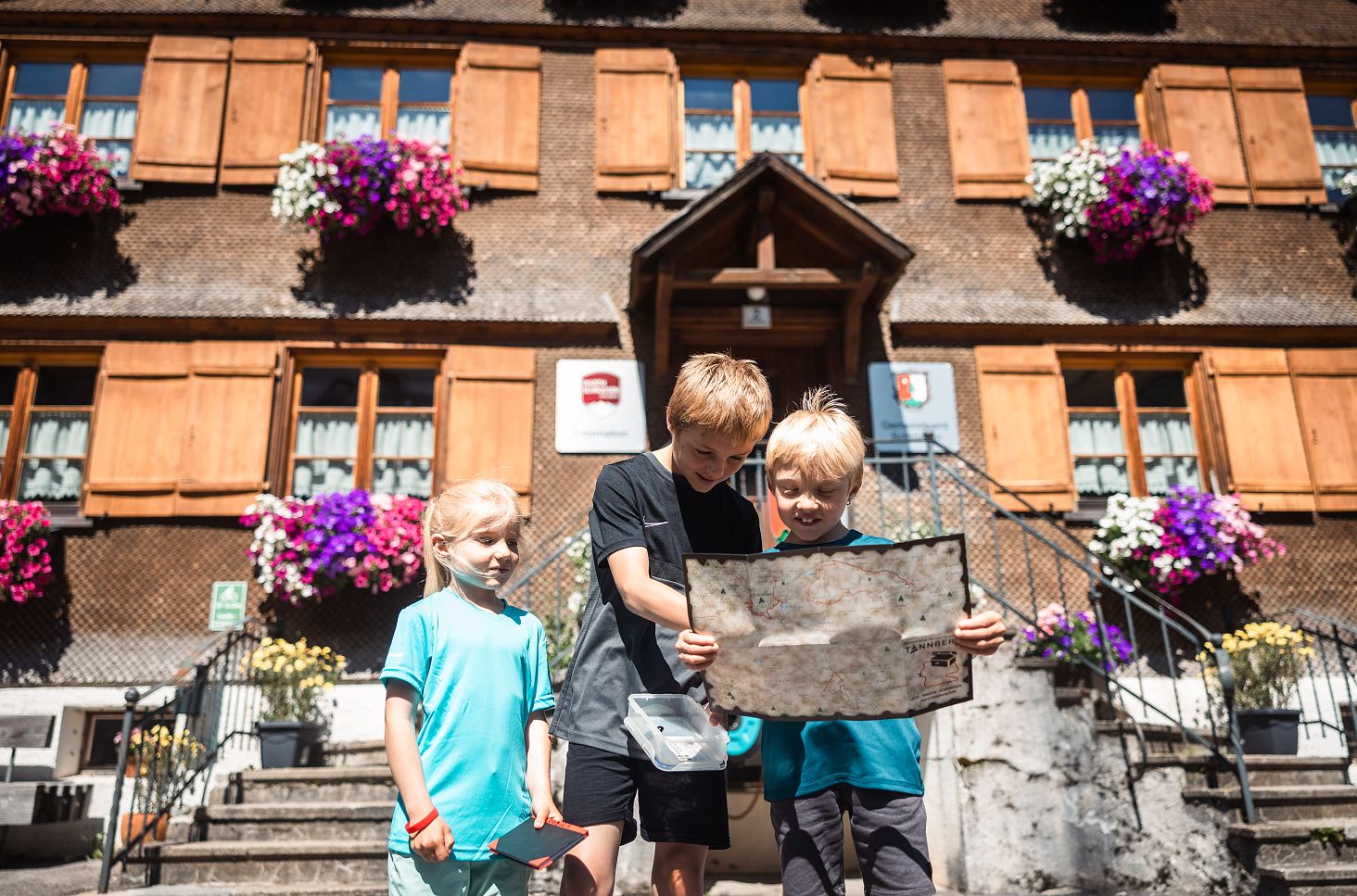Three children stand in front of a traditional wooden house adorned with colorful flowers. They study a treasure map intently as they prepare for their geocaching adventure.