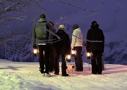 Eine Gruppe von Menschen mit Laternen wandert bei Dämmerung durch eine verschneite Winterlandschaft.
