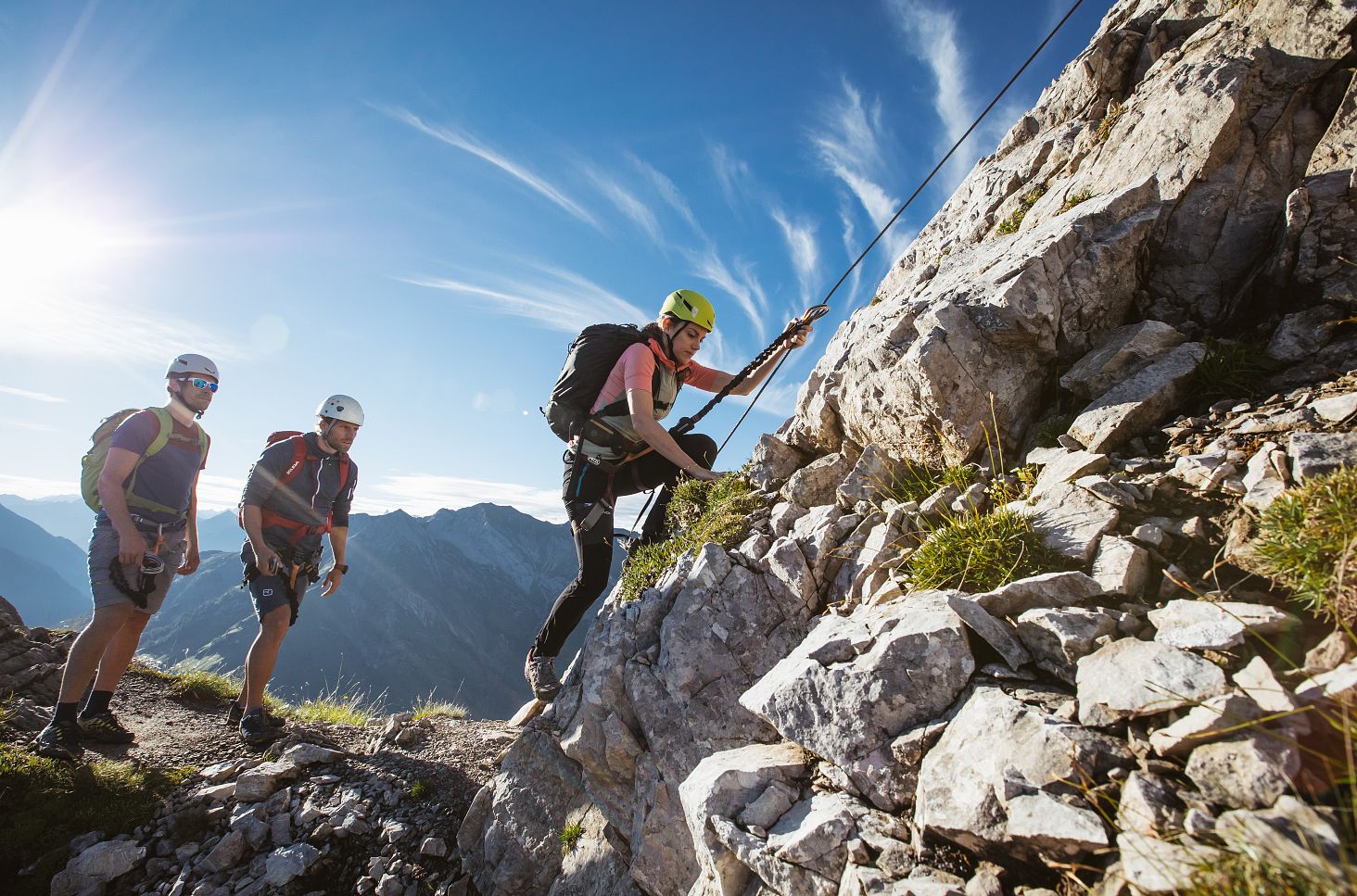 Three climbers navigate the Karhorn Via Ferrata, with a bright blue sky stretching out behind them.