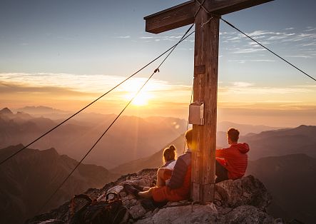 As the sun rises behind the Widderstein summit cross, bathing the mountains in golden light, two people watch in awe.