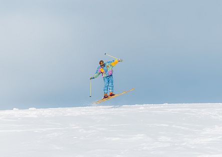 A man in a colourful 1980s ski suit is jumping into the soft spring snow.