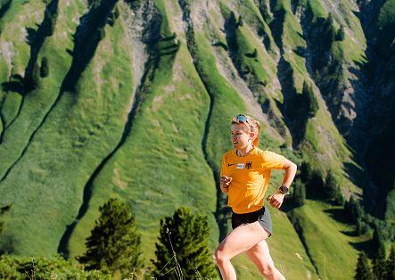 A female trail runner in a yellow shirt is out in the mountains, with a mountain landscape in the background.