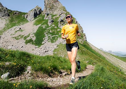 A female trail runner in a yellow shirt is running towards the Wartherhorn, with a mountain landscape in the background.
