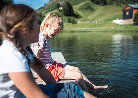 Two children sit on the shore of Seebachsee, dangling their feet in the water.