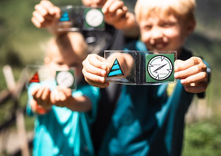 Zwei Kinder halten stolz eine Geocaching-Marke in die Kamera, während im Hintergrund die beeindruckende Berglandschaft von Schröcken zu sehen ist.