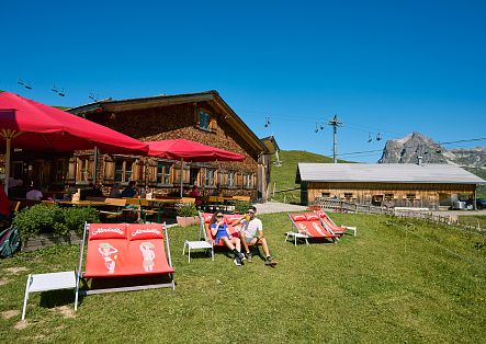 Zwei Personen sitzen entspannt in Liegestühlen vor der Hochalphütte auf einer blühenden Wiese.