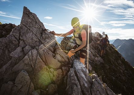 Ein Klettersteigabenteuer in den Bergen: Die Sonne strahlt durch die Felsen, während ein Kletterer mit Helm und Klettergurt konzentriert den gesicherten Grat erklimmt.