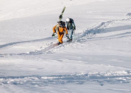 Zwei Personen laufen mit Skiern auf dem Rücken einen schneebedeckten Hang hinauf.