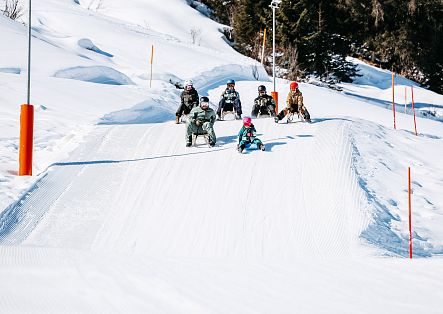 Eine Gruppe Menschen fährt fröhlich mit Schlitten an der Dorfbahn hinunter.