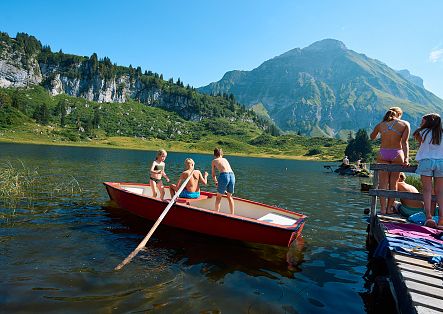 Drei Kinder sitzen in einem Ruderboot auf dem Körbersee, während die restliche Familie am Steg steht.