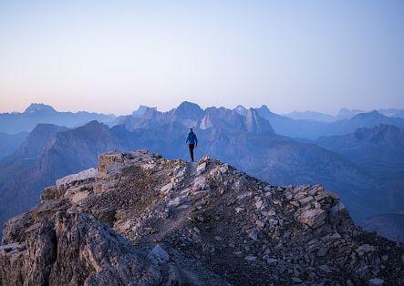 A person stands on a rocky mountain peak, surrounded by a stunning alpine panorama in soft twilight.