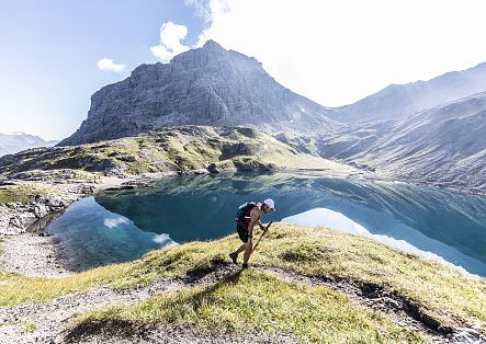 Ein Wanderer erklimmt eine grüne Anhöhe vor der atemberaubenden Kulisse eines kristallklaren Bergsees und einer imposanten Felslandschaft.