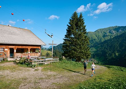 Two hikers walking past an alpine hut, which offers a wide view over Schröcken.