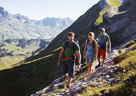 Three people are hiking on a rocky path, with a stunning view of the landscape unfolding around them.