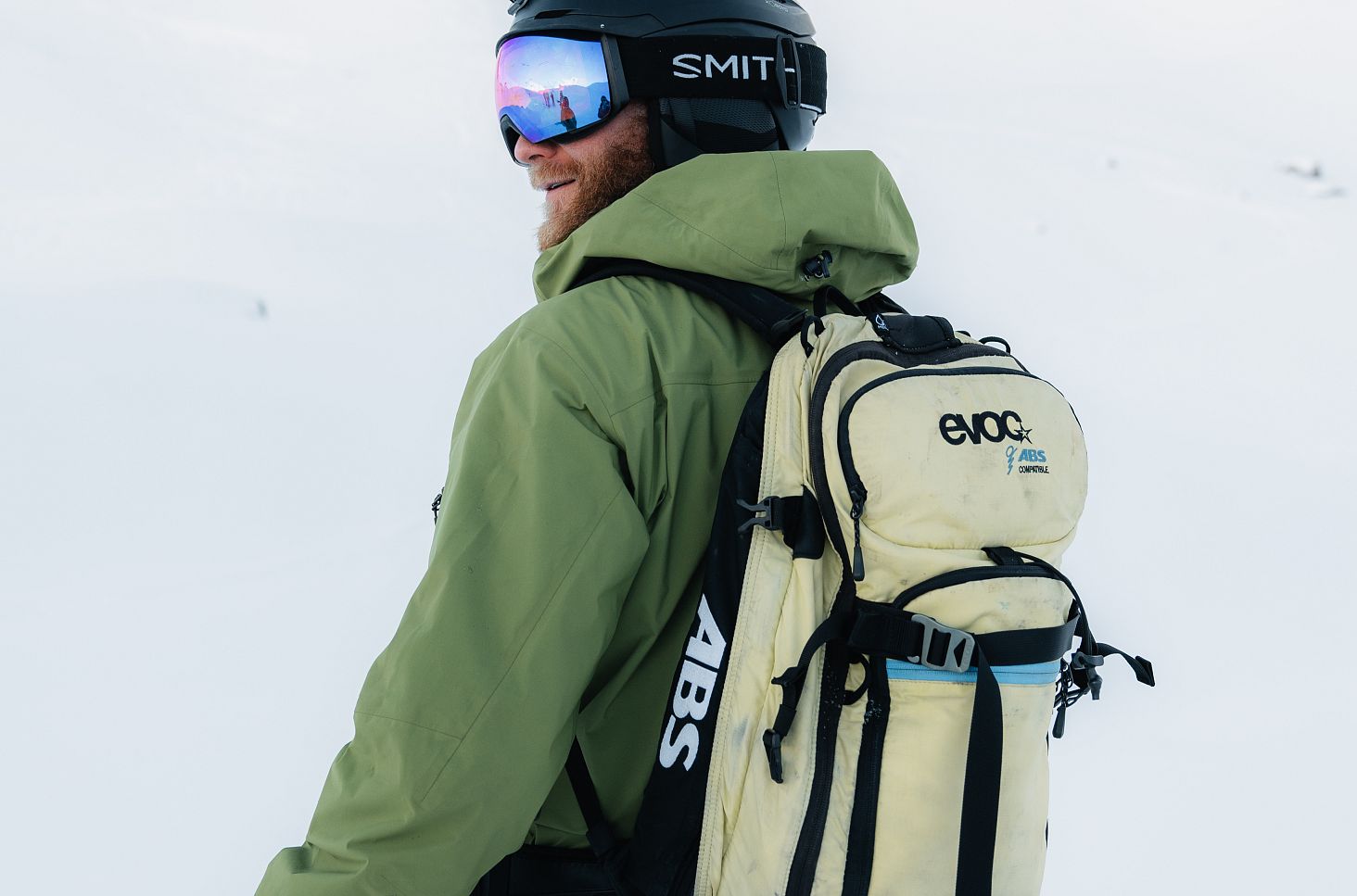 Ein Herr von hinten in grüner Jacke steht im frischen Pulverschnee und trägt einen Lawinenrucksack.