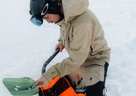 Ein Skifahrer in heller Kleidung beugt sich über seine Skiausrüstung und hantiert an einem Ausrüstungsgegenstand.
