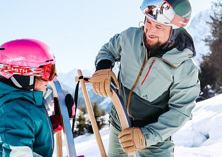 Ein Erwachsener unterhält sich mit einem Kind, das neben einem Schlitten im Schnee steht.
