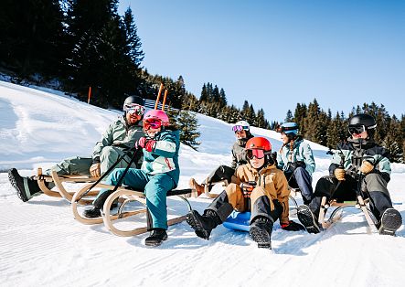 Mehrere Erwachsene und Kinder sitzen gemeinsam auf Schlitten auf einer verschneiten Rodelbahn.