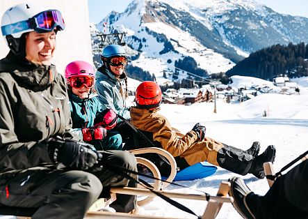 Eine Familie sitzt auf mehreren Schlitten und genießt vor verschneiten Bergen eine Rodelpause.
