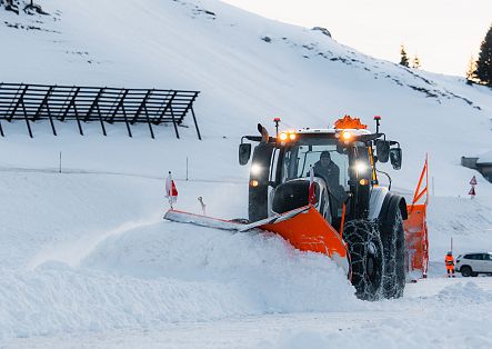 Ein Traktor mit Schneepflug schiebt eine große Menge Schnee auf einer verschneiten Bergstraße zur Seite.