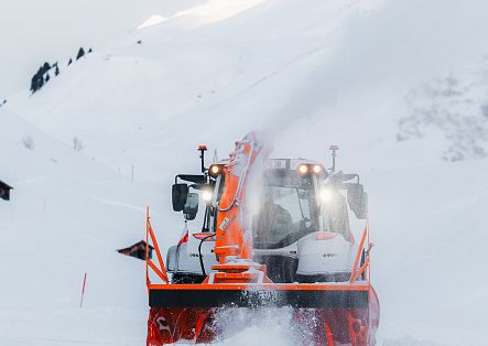 Eine große Schneefräse fährt durch eine verschneite Berglandschaft und wirft Schnee zur Seite.