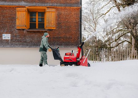 Eine Person räumt mit einer motorisierten Schneefräse den tief verschneiten Bereich vor einem Holzhaus.