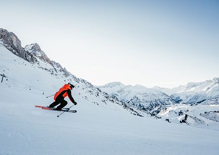 Ein Skifahrer fährt über eine weite verschneite Piste vor einer alpinen Bergkulisse.