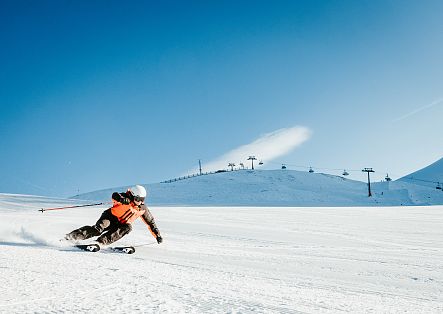 A skier in an orange suit is hurtling down a snow-covered slope, leaning forward deeply in a dynamic posture.