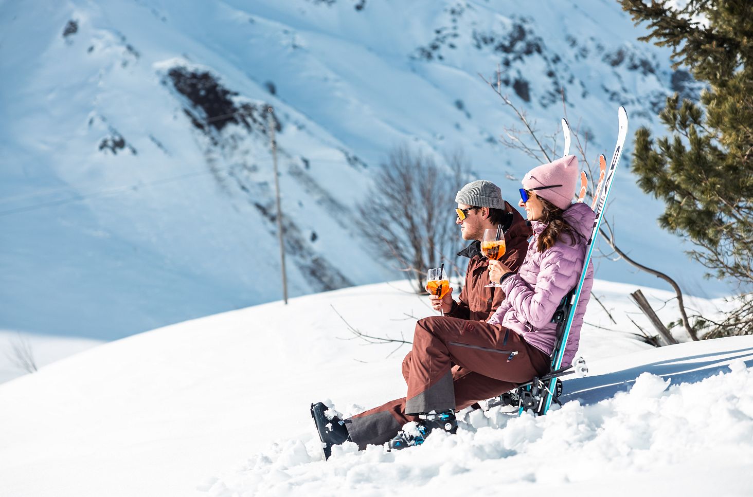 Two skiers make themselves comfortable on their skis and toast with an Aperol in the sun.