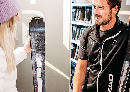 Ein Gast und ein Mitarbeiter betrachten gemeinsam einen Ski in einem Sportgeschäft.