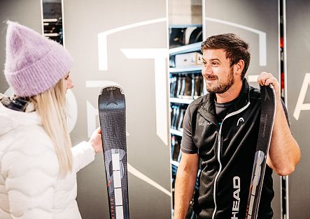 Zwei Personen vergleichen in einem Sportgeschäft einen Ski und eine Skiausrüstung.