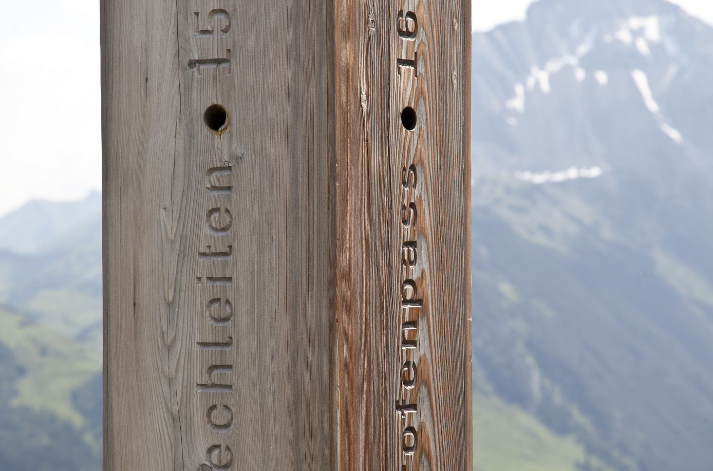 Eine Holzsäule mit eingravierten Ortsnamen und Höhenangaben vor einer alpinen Bergkulisse.