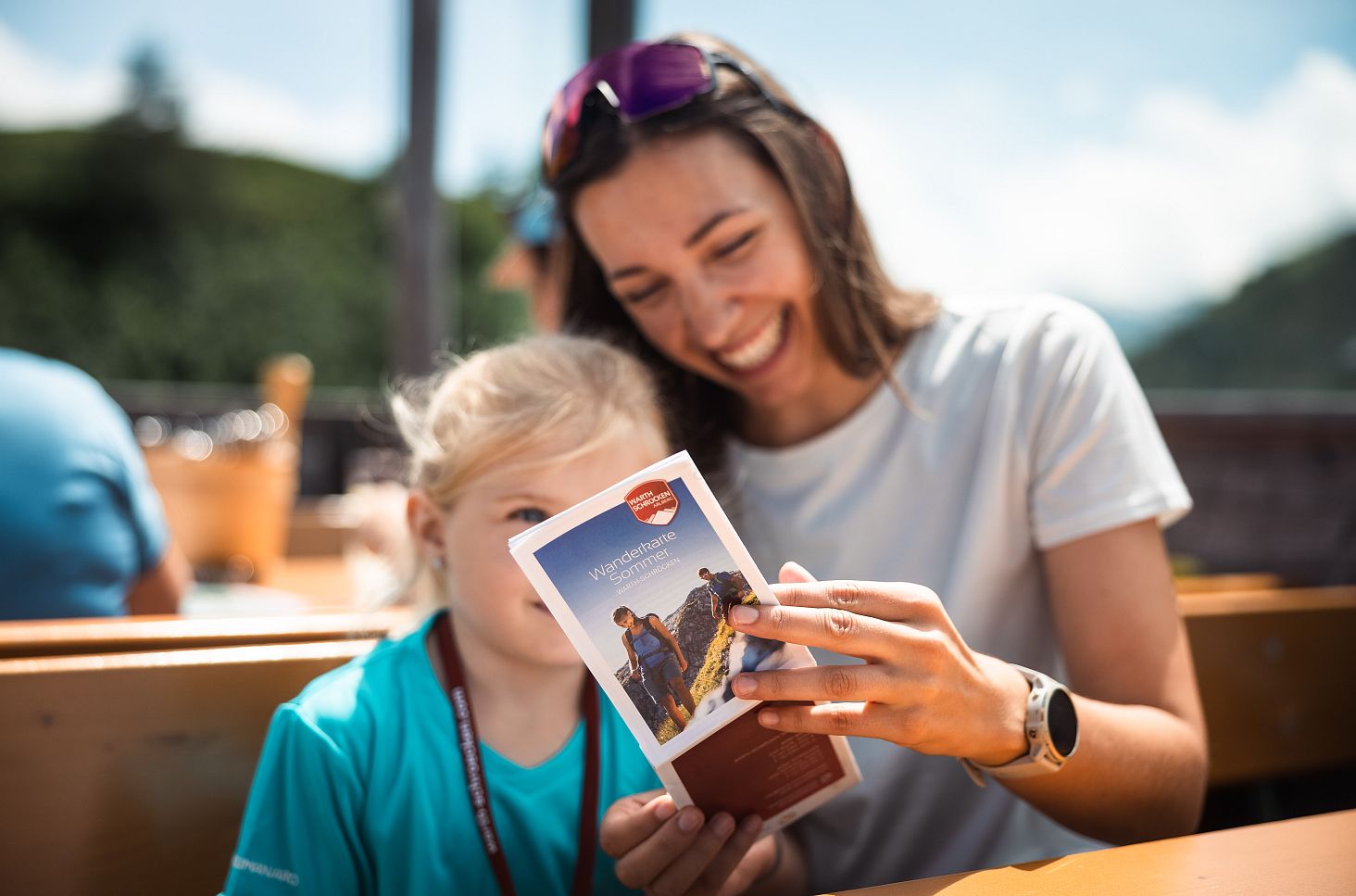 A smiling woman and a child look at a hiking brochure together while sitting at a wooden table outdoors.